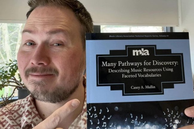  A man with short brown hair and a beard is holding a book in front of his face. He is smiling and pointing at the book with his right hand. The book is titled "Many Pathways for Discovery: Describing Music Resources Using Faceted Vocabularies" by Casey A. Mullin.