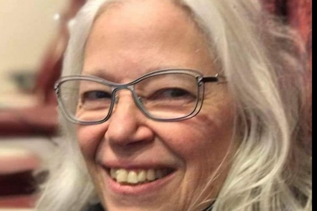 The photo Joann with gray hair and glasses smiling. She is wearing a black shirt with a gray and black striped scarf. There are books and awards on the shelf behind her.