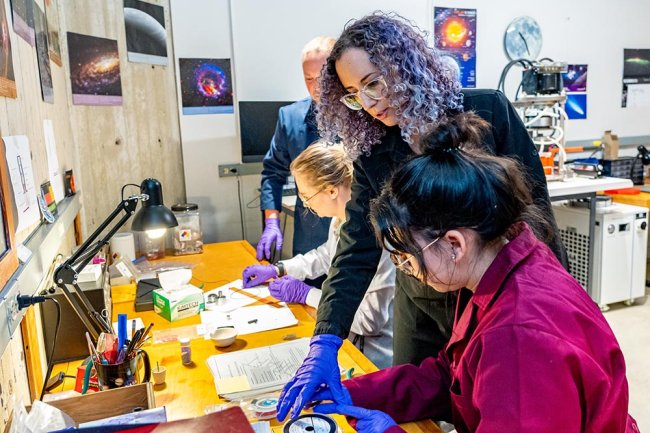 In this image, there are four people in a laboratory setting. On the left, there is a person with shoulder-length brown hair wearing a white lab coat and blue gloves. They are holding a pair of tweezers and appear to be working on a small object. In the middle, there is a person with curly purple hair wearing glasses, a black shirt, and blue gloves. They are looking at something on the table. On the right, there is a person with long black hair wearing a maroon lab coat and blue gloves. 