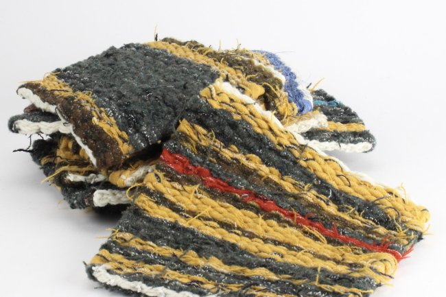 The image shows a handwoven rag rug made of recycled materials. The rug has a striped pattern in brown, yellow, blue, and red. The edges of the rug are frayed and uneven. The rug is lying on a white background.