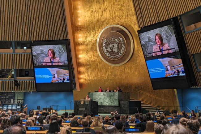  In the United Nations General Assembly Hall, a woman stands at a podium, speaking into a microphone. She is wearing a suit and has short brown hair. There is an emblem on the wall behind her. The hall is filled with people, mostly young people, who are sitting in rows of chairs. They are listening to the woman speak. On either side of the woman there are two large screens which show a close up of her face.