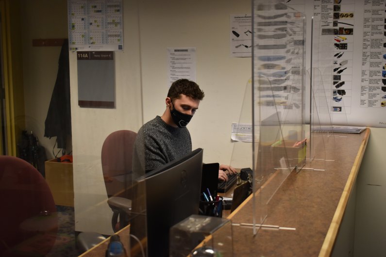 A masked student works at a check-in desk behind a plastic divider.
