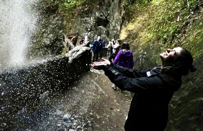 A student holds out their hands to catch the spray from the powerful Pailon del Diablo waterfall as they stand on a nearby walkway.