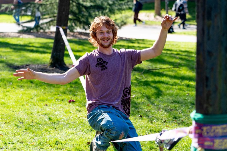A bearded, red-haired student balances on a slack line between two trees on campus.