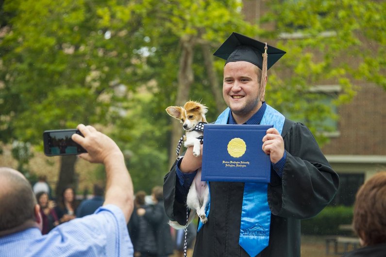 a graduate smiles for a photo while holding a diploma cover and a small dog