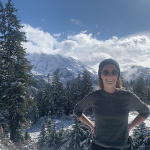 Olana Costa on a snowy mountain ridge on a sunny winter day; behind her are a set of snowy peaks