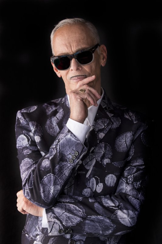 John Waters poses with his hand to his chin wearing sunglasses.