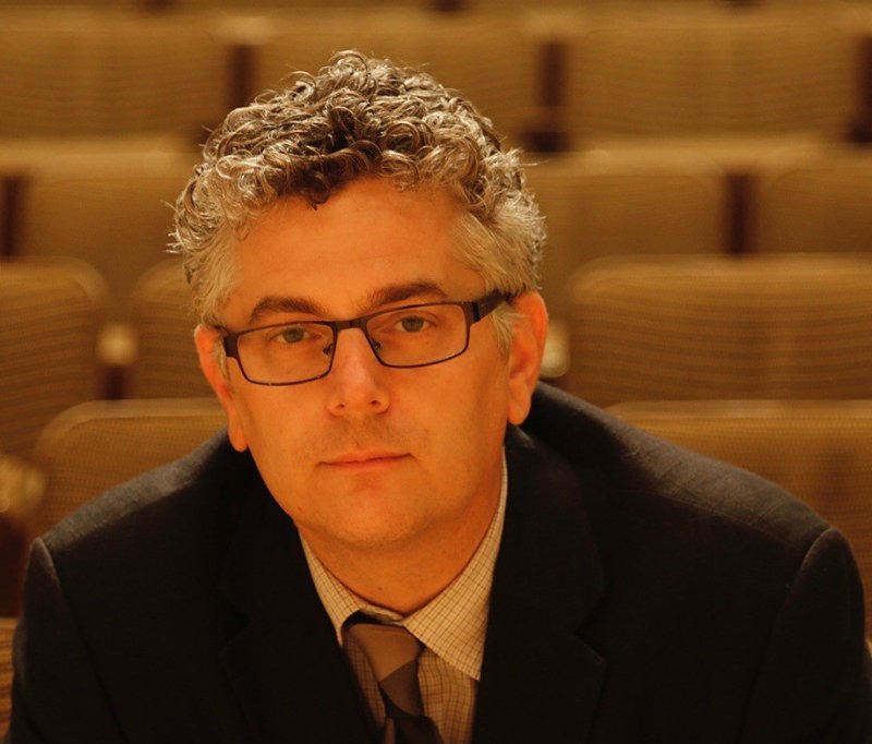 Chris Bianco looks at the camera solemnly; behind him are the seats from a concert hall