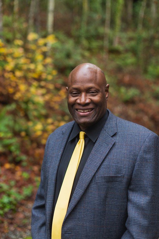 Leonard wears a yellow tie with dark blue suit jacket, and poses near the Sehome arboretum