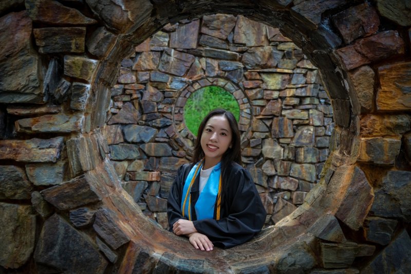 Aika Okayama smiles, wearing academic regalia, including a sash and honors cords, while surrounded by one of the circular openings in Stone Enclosure: Rock Rings sculpture