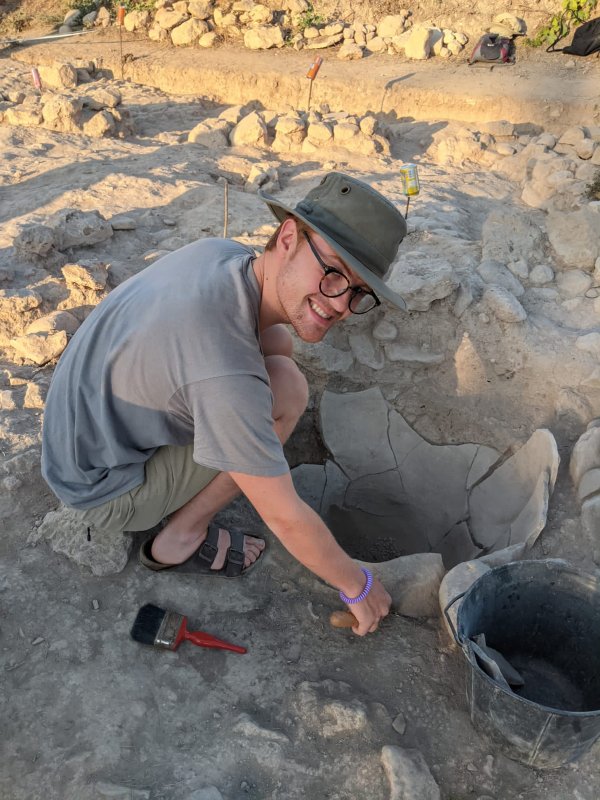 Jack Tillman crouches over a hole in the midst of an archeological dig, bucket and dust brush within hand's reach. He turns and smiles for the camera
