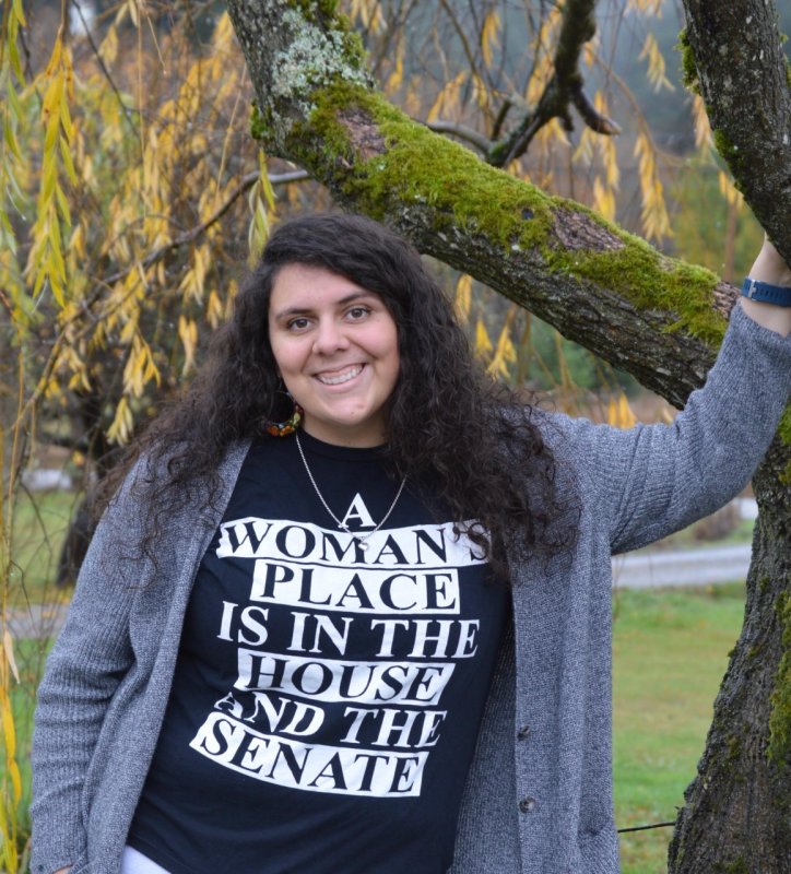 Lindsay Little wears a shirt that says “A Woman’s Place is in the House and the Senate” as she leans against a tree. 