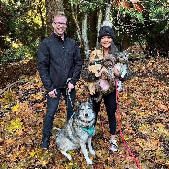 three people stand outside in a wooded setting. One holds two small dogs and a third larger dog sits in front of the people