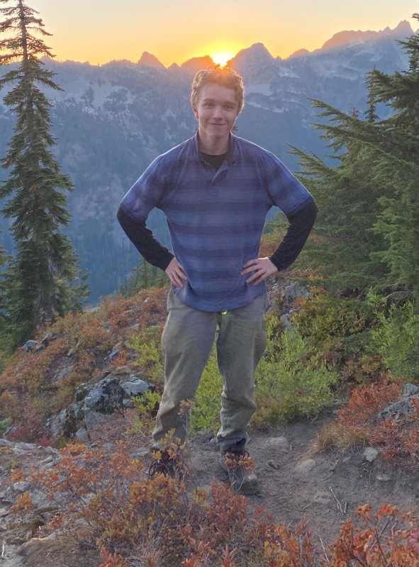 Nick Satnik stands, smiling, on a mountainside, the sun setting in the background