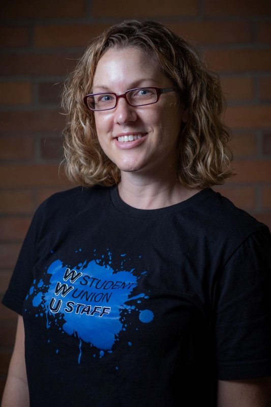Jenn Cook wears a WWU Student Union Staff shirt and smiles in front of a brick background.