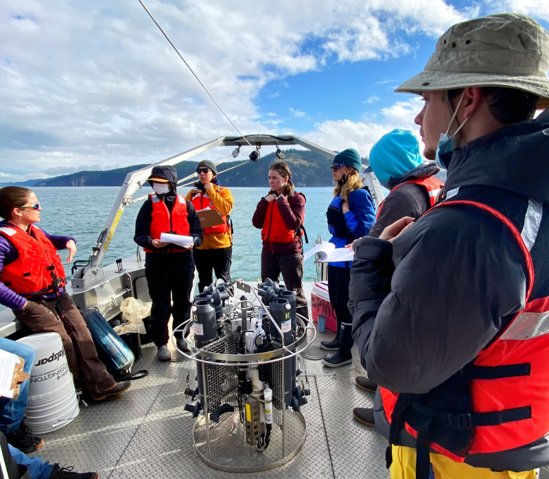 Students listen to an instructor at the stern of Western's research vessel; behind them are blue skies and one of the San Juan islands