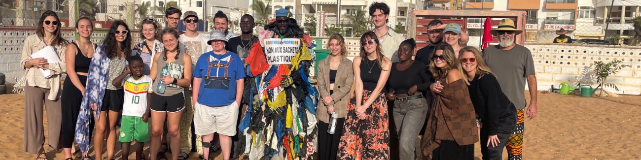 Students and faculty stand on a beach in Senegal, with a man wearing dozens of plastic bags attached to his clothing and a sign saying 'No Plastic Bags," in French.