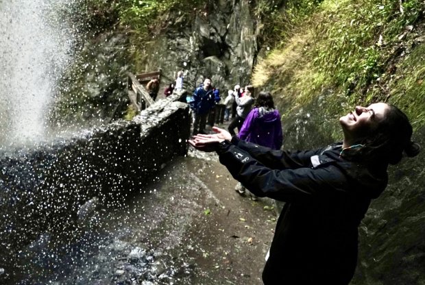 A student holds out their hands to catch the spray from the powerful Pailon del Diablo waterfall as they stand on a nearby walkway.
