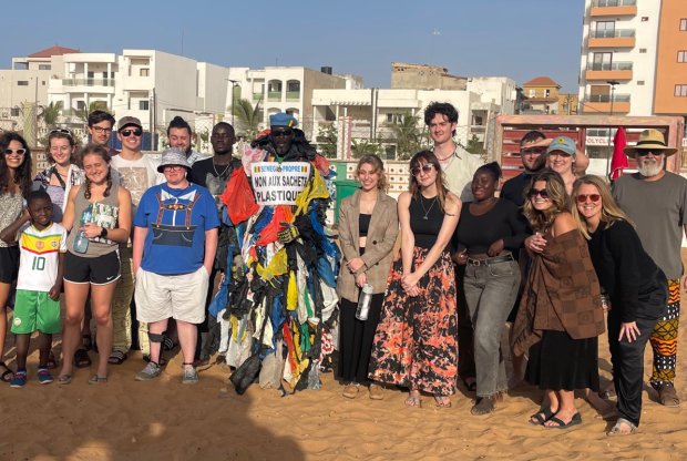 Students and faculty stand on a beach in Senegal, with a man wearing dozens of plastic bags attached to his clothing and a sign saying 'No Plastic Bags," in French.