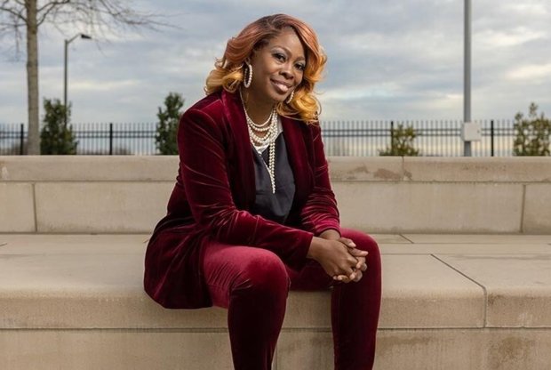 LaTosha Brown sits on a marble step and smiles at the camera; behind her are clouds