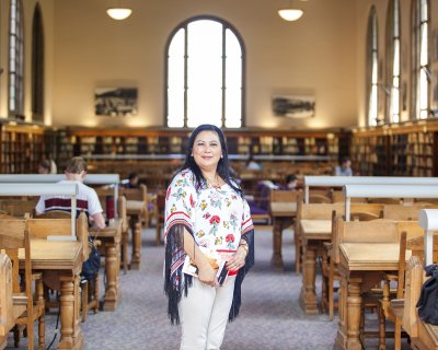 Blanca Granados de Cruz facing the camera while standing in the Wilson Library Reading Room holding a book 