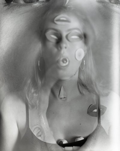 Black and white portrait of Emily Keogh facing directly towards the camera with stickers of lips on their forehead, eyes and cheeks photo is also slightly blurred