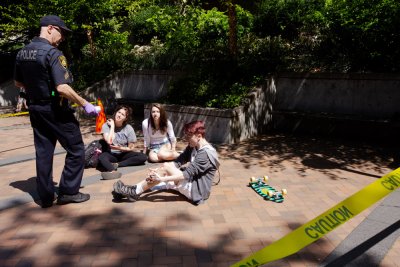 A police officer talks to students who are sitting on the ground, surrounded by yellow caution tape, following a mock accident to illustrate the importance of pedestrian safety