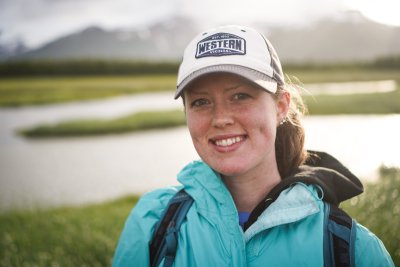 Juliana Covarrubias smiles, wearing a backpack and a WWU hat, with a mountain and river in the background