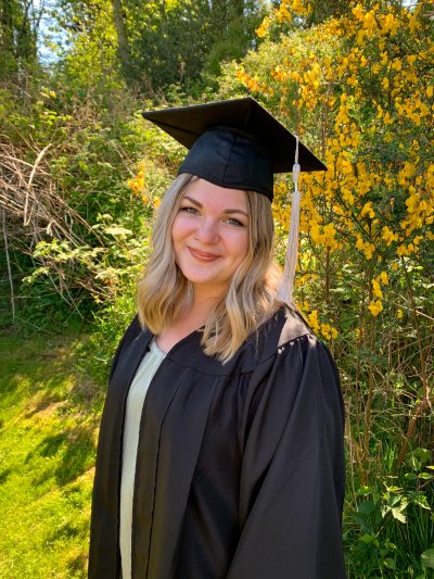 Rylee Tuttle smiles while wearing graduation regalia in front of a flowering tree
