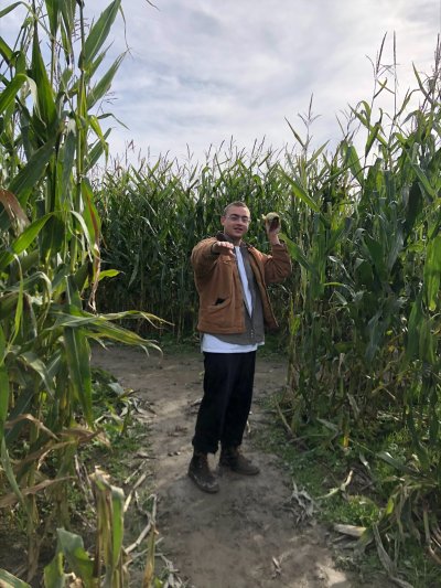 Tower Bert stands in a corn field holding a corncob above his shoulder with one hand and reaching out toward the camera with his other hand, as if he were preparing to throw a football