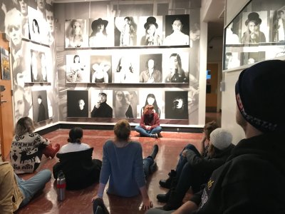 Students sit in the center of the Big Heads exhibition 5 students are sitting on the floor and one is standing
