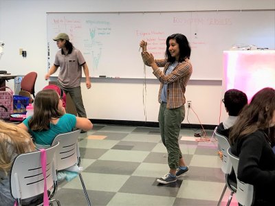 stands in front of a classroom of children, holding string and demonstrating a concept